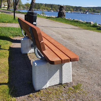 Eine hölzerne Parkbank mit Rückenlehne auf einem Betonrahmen, die an einem Spazierweg an einem See steht.